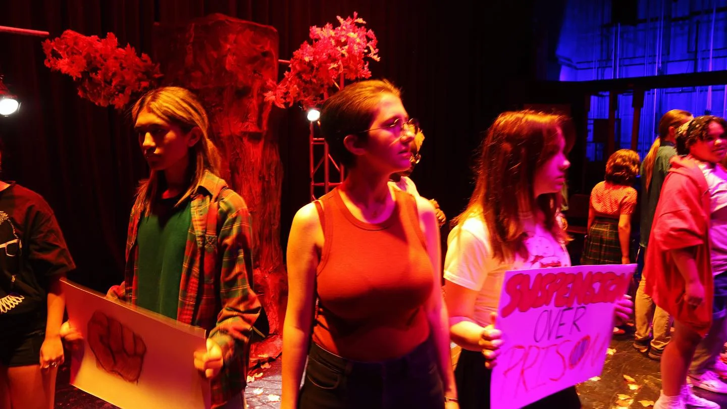 The Blood at the Root ensemble marches across the stage carrying protest signs under red and blue lighting.