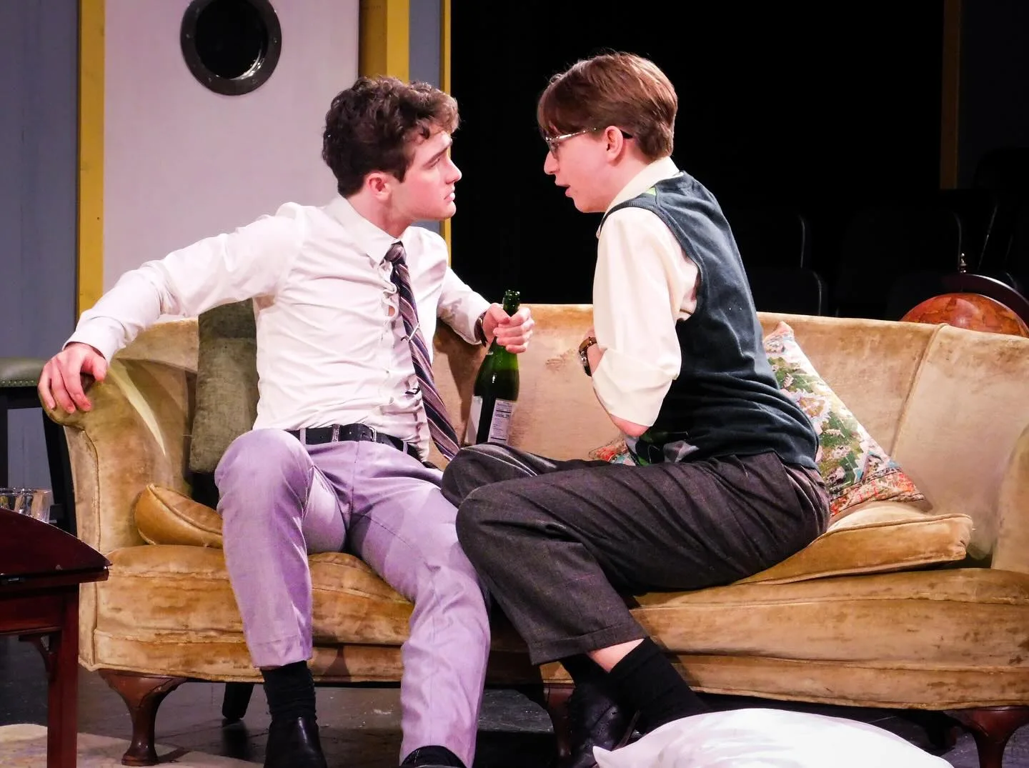 Two Boeing Boeing cast members confer on a sofa in Bernard's Paris apartment, one holding a champagne bottle.