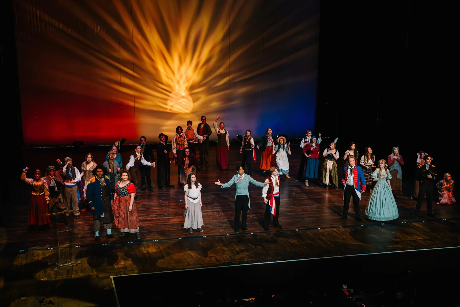The Les Misérables company performs onstage beneath red, gold, and blue lighting.