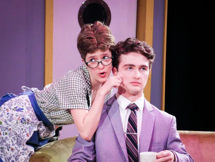 Two student actors perform a comic scene on a living-room set.